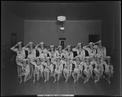 Juanita Crutcher; Dance Group at Mac. Hall, girls dressed in outfits with hats on lined up in two rows saluting, interior of empty room