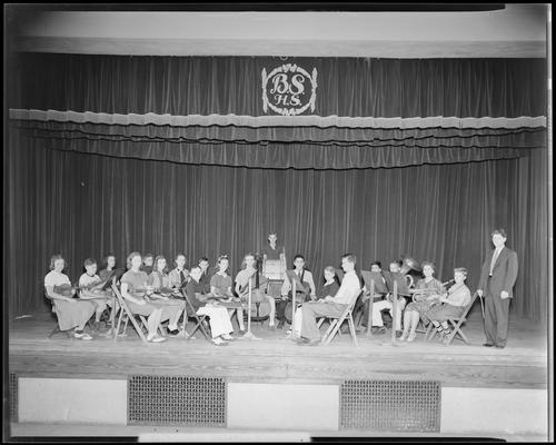 Bryan Station High School; orchestra group portrait, orchestra members (students) sitting on stage with their instruments, conductor standing to the side