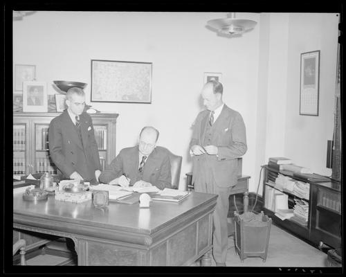 Kentucky Utilities Company (167 West Main Street); P.S. Wester, R.M. Walt, & A. Tuttle; interior of office, three men gathered around a desk, one of the men is sitting at the desk signing a document
