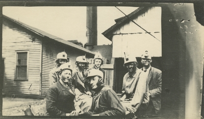 7 men and women (appear to be the same people as item #6 and #7) at a coal camp with buildings in the background