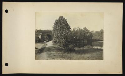 Dirt roadway on left of picture leading under stone arch of bridge, river or pond on right with metal fenced in area
