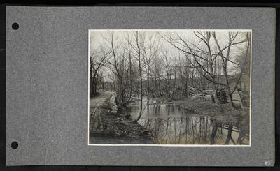 Small river with two ducks swimming in distance, roadway on left, wooden fencing on right