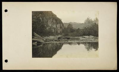 Calm river with sizeable rocks across river and on banks, bluffs on left bank, buildings in extreme distance on hilltop