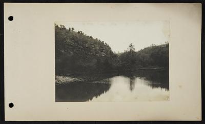 River with riffles and floating debris, bluffs rising on left side of picture