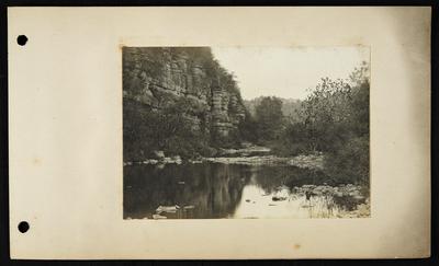 Calm river with flat rocks on right bank extending almost to left bank, bluffs on left, buildings on hilltop in distance