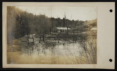 Possibly flooded river running in front of vaguely Misson Style building, two round buildings in front, round structure connected to back of front building, railed walkway running up hill, low building to far right, possibly a house behind front building, buildings on hilltop in distance