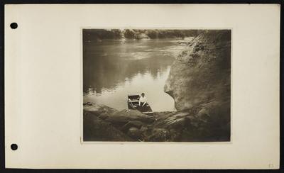 Woman sitting in boat with one paddle, large river behind her, large rounded rock to right