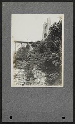 View of bridge leading off of cliff, man standing on top of bridge, large stone archway, bluffs below