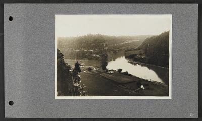 River running into a fork, timber along banks, collection of buildings on far right bank, houses on left bank, small structure on top of bluff with steep stairs