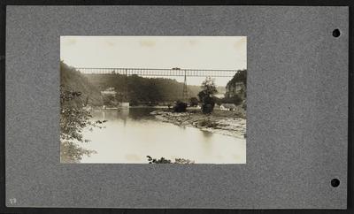Railroad bridge spanning over river, engine and car traveling along, timber along banks of river with two houses, two boats on river