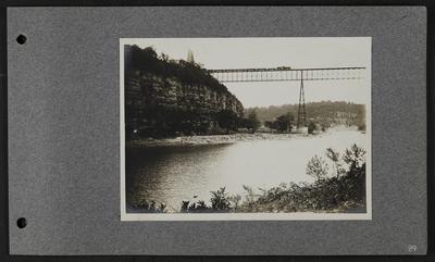 Railroad bridge over river with train traveling along, lots of timber along far river bank, two women and one man walking along far bank