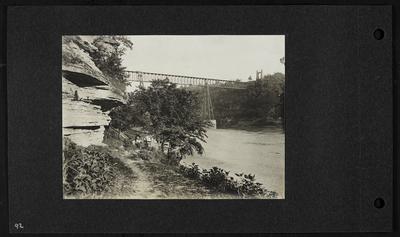 Five people on walkway along river with bridge in background, broken fence along path