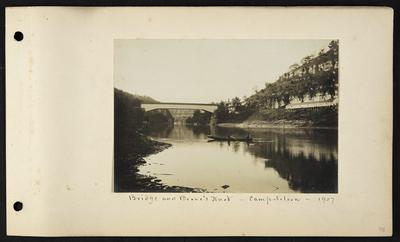 Two men in boat rowing along river, bridge in distance, houses near bridge, long low building on right, notation                          Bridge and Boone's Knob, Camp Nelson, 1907