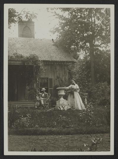 Lady standing at urn, man and child seated in a yard in front of a house