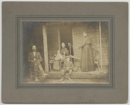 Unidentified group sitting on a porch- one man, two children, and two women. The back of the photograph reads: How Mountain Women spend their leisure hours. Mountain women spinning