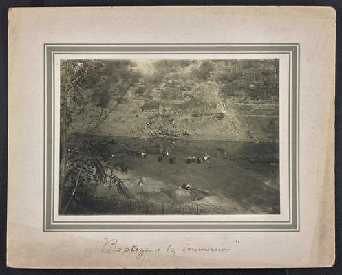 A large group of unidentified people standing around a river bank with a cabin nearby. The bottom of the photograph reads: Baptizing by immersion