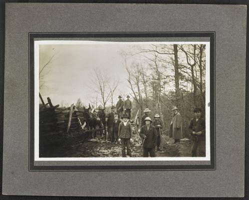 Unidentified men and young boys standing on a dirty road with tools, horse buggy in the back. The back of the photograph reads: Building model road at Cranston, Marion Ross, Road Engineer, Slate Department Roads