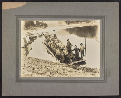 Seven unidentified males on a push boat, packed with supplies, pulled to shore. Back of the photograph reads: Conveying supplies up East KY streams before railroad was built