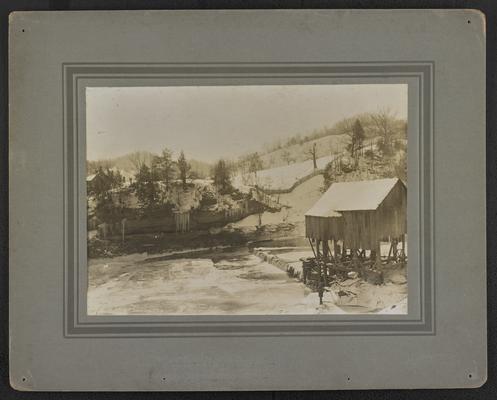 A log cabin sitting by a frozen creek. The back of the photograph reads: The Mountain Child- His Environment. His isolation even hath its charms