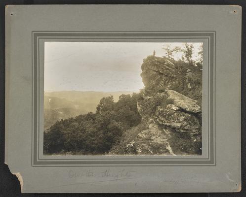Unidentified man standing on a mountain cliff, overlooking mountains and trees. Below the photograph: On the Heights. On the back of the photograph: The Mountain Child