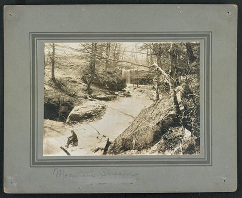 Unidentified man sitting by a stream in the woods, waterfall in the background. Below the picture reads: Mountain Stream. On the back of the photograph: The Mountain Child