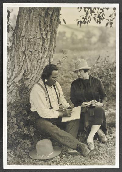 Cora Wilson Stewart sitting with a Native American male at the base of a tree, while he writes in a notepad and she looks on