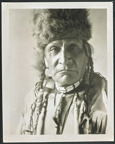 Formal portrait of a man wearing traditional Native American dress. Back of the photograph identifies him as: Chief Curly Bear, a venerable Blackfoot Indian