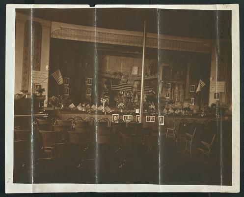 A stage decorated with portraits of the same man and American flags. The back of the photograph reads: The opera house at Marion, Kentucky decorated for my speech there in October 1920 signed by Cora Wilson Stewart