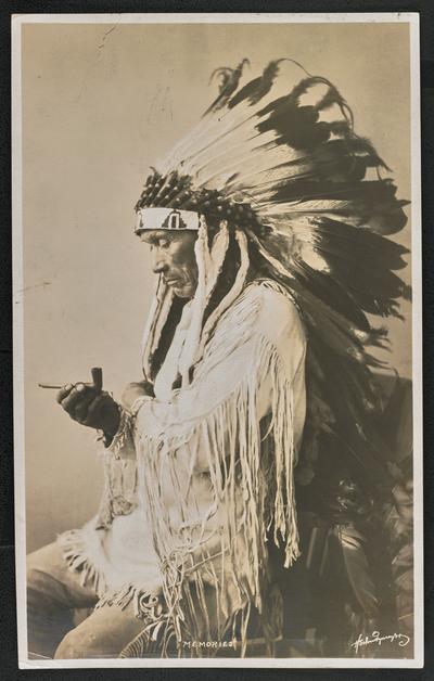 Formal portrait of a man wearing traditional Native American dress. The back of the photograph identifies him as: Koosta. Chief of the Kootena: Indians, Palson, Montana. Koostata is 68 years of age. Son of the famous Chief Eneas, and grandson of chief Bapsiste, who signed the treaty of Hell Gate, 1855, between the U.S. Government and the Allied Tribes of the Flashed Reservation