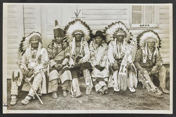 Six unidentified males sitting and wearing traditional Native American dress, four of them wearing headdresses