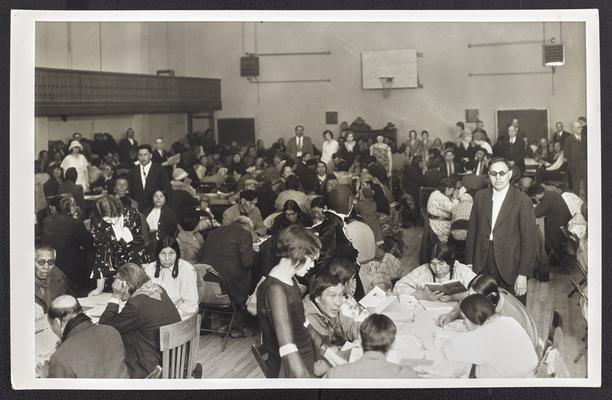 Native Americans sitting around tablse reading books in a large room at the Blackfeet Gymnasium