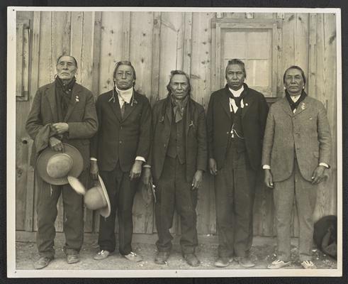 5 males standing outside. Back of the photograph reads: Indian Speakers reading left to right: Black Weasel, Two Guns White Calf, Judge No Coat, Charlie Iron Breast, and Yellow Kidney. Illiteracy Clinic, Blackfeet Indian Reservation