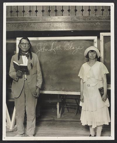 Native American man and his teacher stranding in front of a chalk board. The back of the photograph reads: Writing Contest -John Little Blaze- Winner. Illiteracy Clinic, Blackfeet Indian Reservation