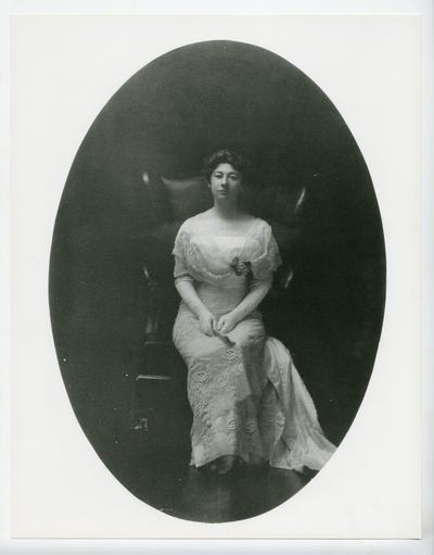 Full-length, formal portrait of Cora Wilson Stewart wearing a white dress and gloves, sitting in a leather chair
