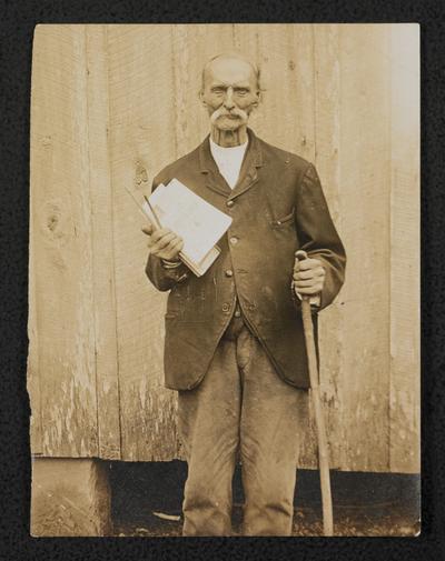 An elderly man standing outside holding books and pencils, back of the photograph reads: A happy school boy; went about teaching
