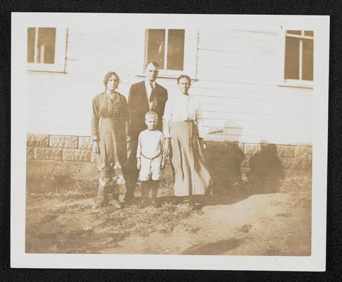 Students standing out front of a school house- 1 male, 2 females, and 1 young boy