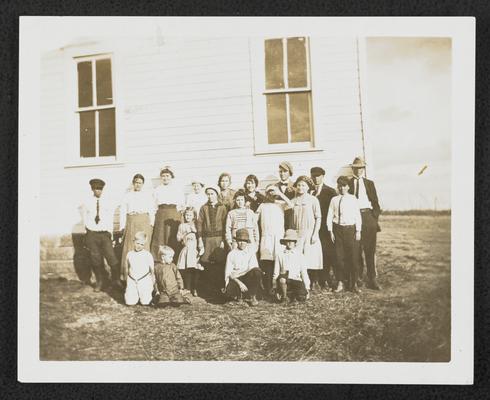 A group of students, all ages, standing in front of a school house
