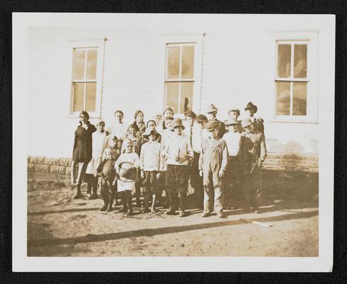 A group of students, all ages, standing in front of a school house
