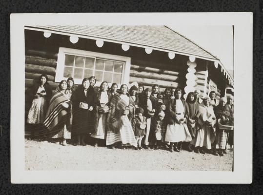 Group of unidentified men, women, and children standing in front of a log building