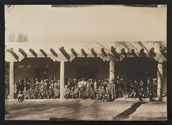 Group, unidentified. Large group of men and women posing on the portico of a Spanish style building