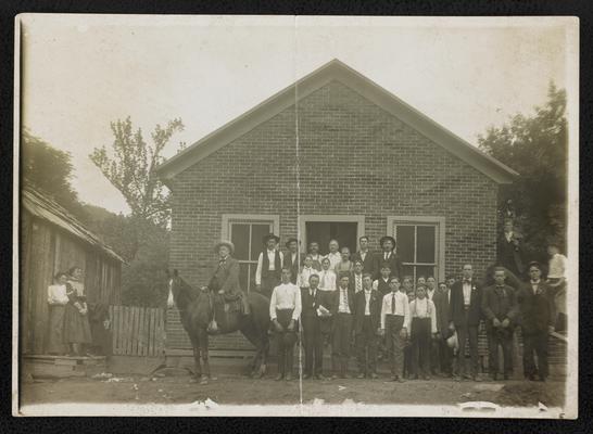 Males, unidentified, standing in front of a brick building. Two women looking of from the side