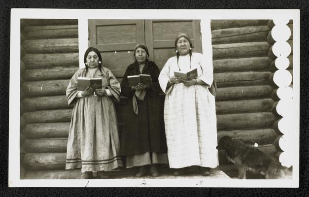 Three women holding books in front a log cabin. Back of the photograph identifies them as: 1. Mary Henry Gun or Hood Looking, age 64 2. Grass Snake, age 52 3. Jane Butterfly, age 54., photograph taken by Richard Sanderville