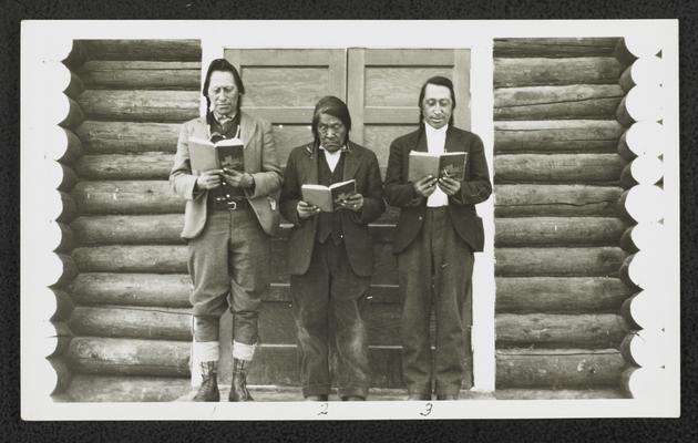Three males holding books in front a log cabin. Back of the photograph identifies them as:  1. Tom Spotted Eagle 2. Red Head 3. James Spotted Eagle