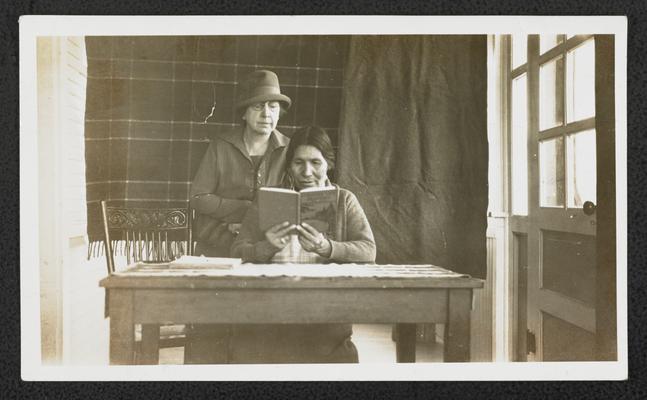 An unidentified white woman looking over the shoulder of a Native American woman while she reads at a desk. Back of the photograph reads: Julia Wades in the Water learning to read