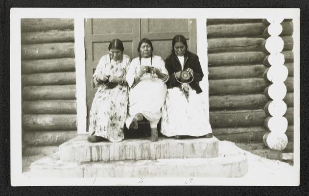 Three women sittign in front a log cabin, sewing. Back of the photograph identifies them as: 1. Mrs. Redfox 2. Strikes First 3. Kills for Nothing