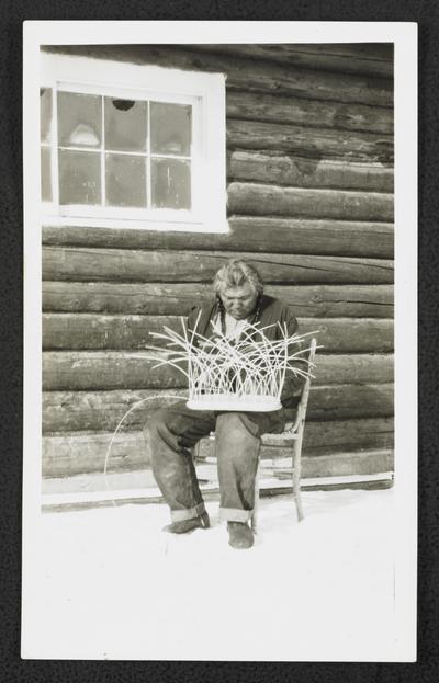 Man sitting outside in a chair, weaving a basket. Back of the photograph identifies him as: Albert Calfrobe