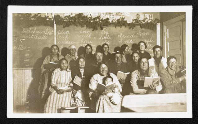 Group, sitting in front a classroom in front of a chalk board, printed in Havre, Montana on February 16, 1927. Paper attached to photograph identifies the students: Top Row: Mary Heavy Gun, Isabelle Jack Spotted Wold, Gretchen Choate, Julia Wades in the Water, Jeanette Lonehorn, Mrs. Orsborne (teacher). Middle Row: Susan Vielle, Medicine Hollering, Calf Rode, Albert Calf Robe, John Spotted Wolf, Philip Surechief, George Lonehorn. Bottom Row: Maggie Spotted Wolf & baby, Henry Heavy Gun, Sam Choate. Bad night and all were not there