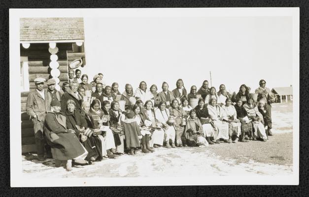 A large group sitting in front of a log cabin, displaying books and handwoven baskets. Back of the photograph reads: Hear Butte Illiteracy