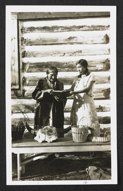 Two women looking at a book, standing behind a display table with woven baskets and an animal skull. Back of the photograph identifies them as: 1. Mrs. Blackbull 2. Irene Butterfly
