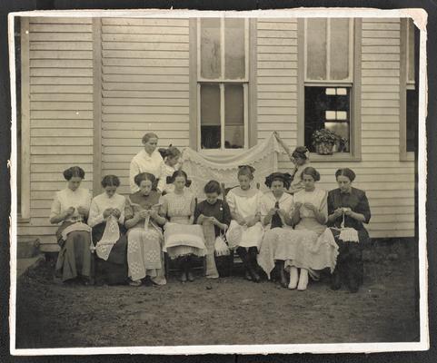 Mrs. C.T. Flannery, teacher. 9 girls sitting and knitting, 2 girls holding a knitted blanket, teacher standing and looking on